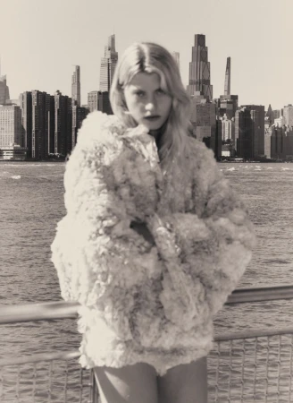 Model wearing a textured white coat standing against a city skyline and waterfront view