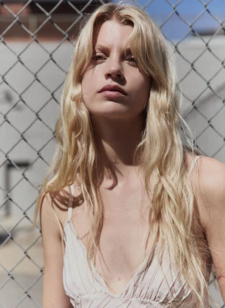 Blonde woman wearing a pale patterned top posing against a chain-link fence outdoors in bright daylight