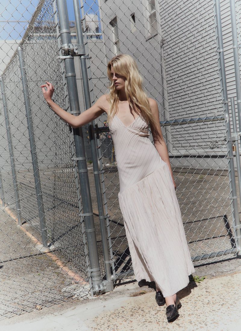Blonde female model in a light-colored slip dress leaning against a chain-link fence outdoors