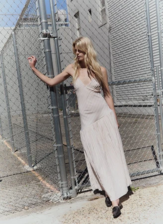 Blonde female model in a light-colored slip dress leaning against a chain-link fence outdoors