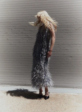 Blonde model wearing a feathered grey dress standing against a textured brick wall with hair blowing.