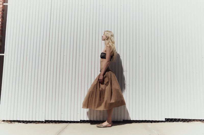 Model Tinka Carleton-Smith in a brown skirt and black top standing against a corrugated metal wall for Modern Road.