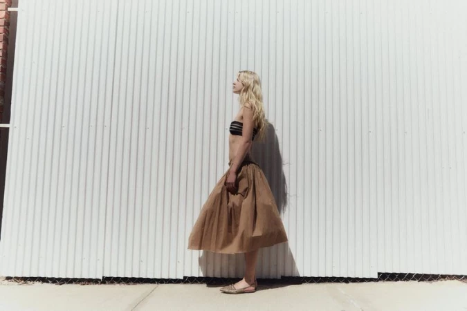 Model Tinka Carleton-Smith in a brown skirt and black top standing against a corrugated metal wall for Modern Road.