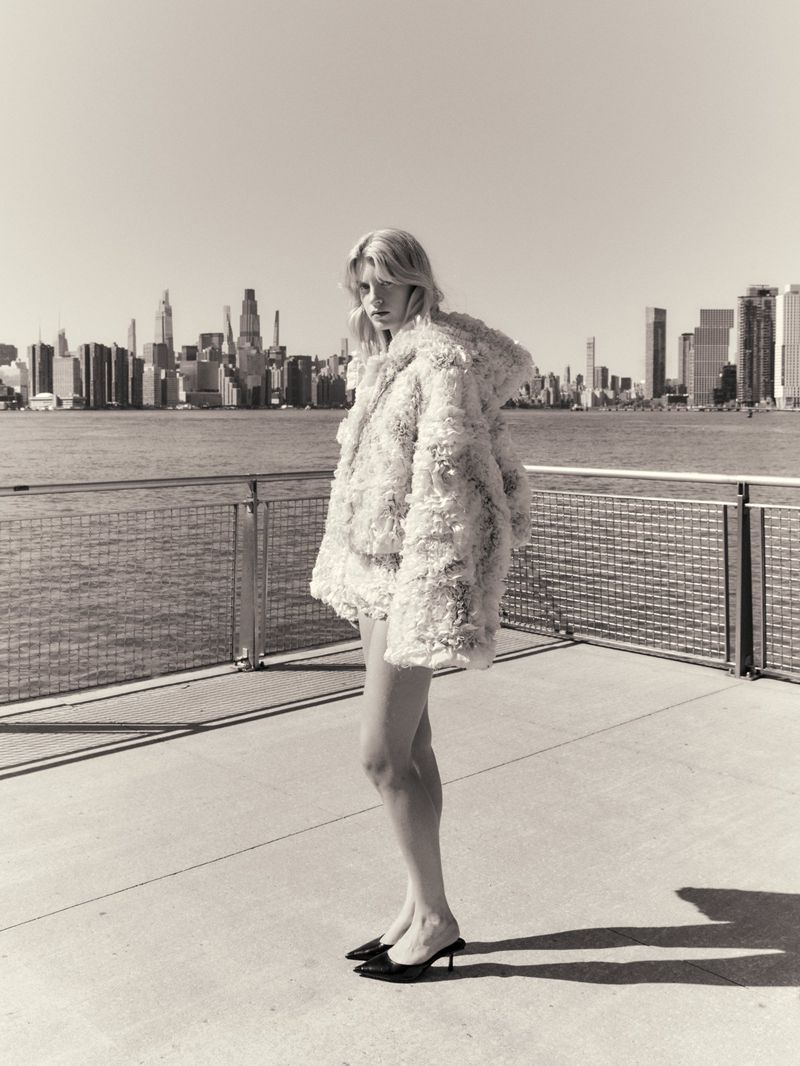 Model standing on a waterfront pier wearing a textured white jacket with a city skyline in the background
