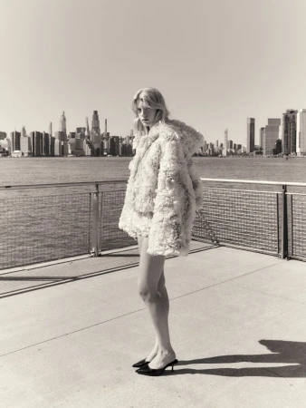 Model standing on a waterfront pier wearing a textured white jacket with a city skyline in the background