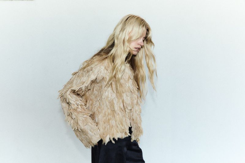 Model with long blonde hair wearing a textured feather jacket against a plain white background