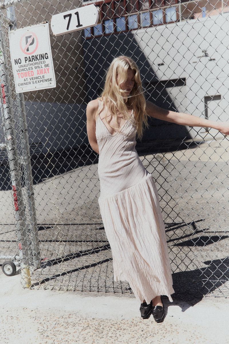 Blonde woman in a cream slip dress leaning against a chain-link fence outdoors