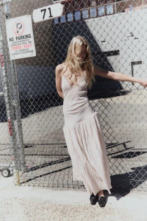 Blonde woman in a cream slip dress leaning against a chain-link fence outdoors