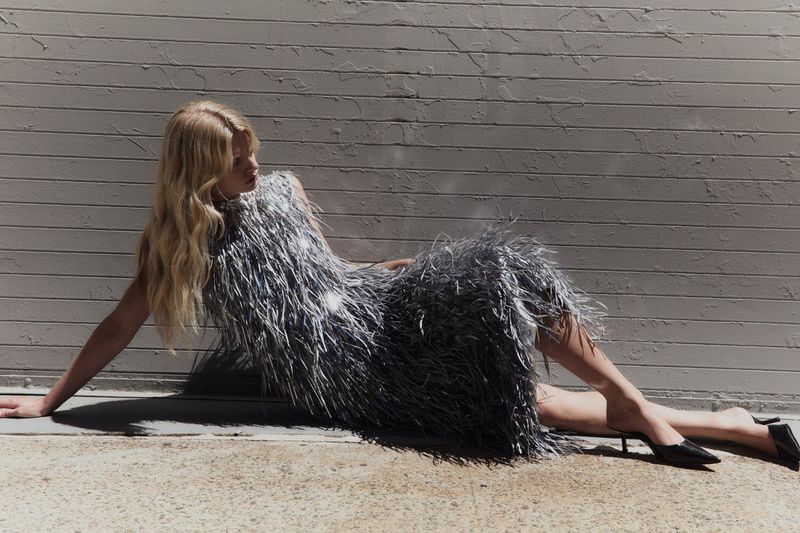 Model in a sparkling silver fringed dress reclining against a textured grey wall outdoors.