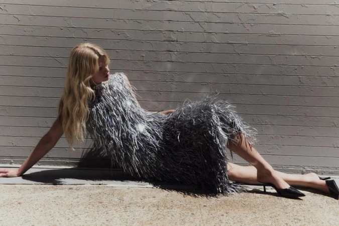 Model in a sparkling silver fringed dress reclining against a textured grey wall outdoors.