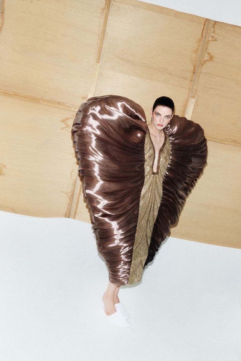Model wearing a structured brown satin sculptural garment against a white and wood panel background for W Korea