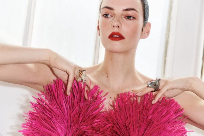 Model wearing a vibrant hot pink textured top and statement rings with red lipstick against a bright white window background