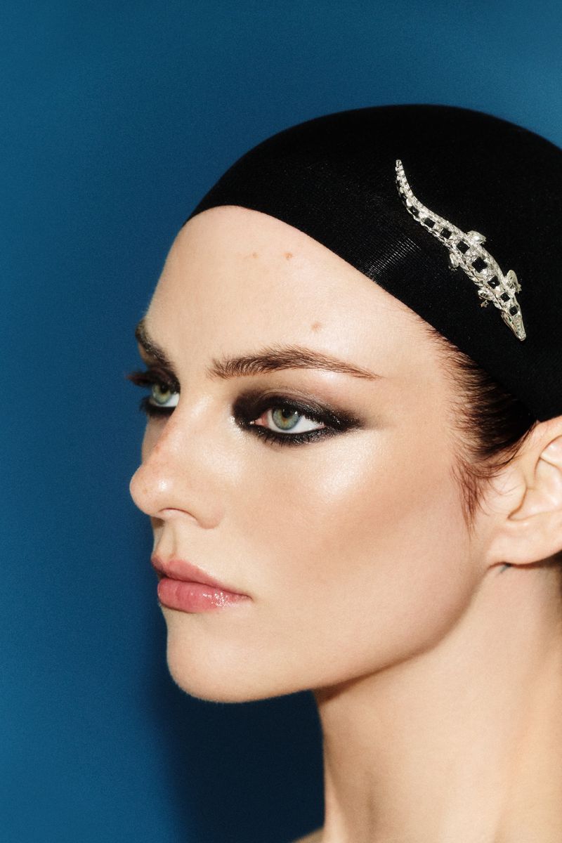 Close-up profile portrait of a woman wearing a black cap with a silver brooch and dramatic dark eye makeup against a blue background