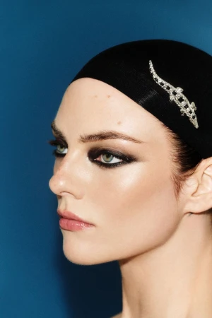 Close-up profile portrait of a woman wearing a black cap with a silver brooch and dramatic dark eye makeup against a blue background