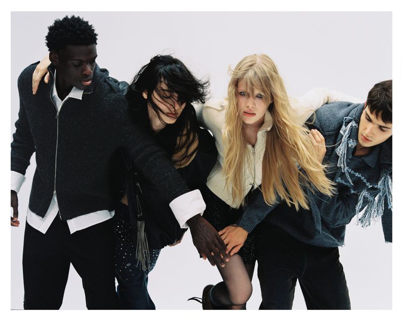 Four models with arms linked posing together against a plain white studio background in an editorial fashion campaign photograph.