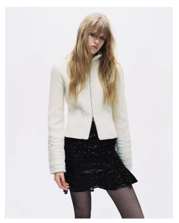Model with long blonde hair wearing a white zip-up cable knit cardigan and black sequin skirt against a white background