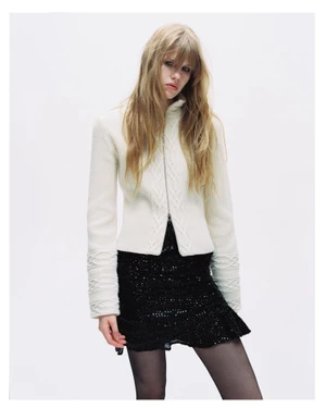 Model with long blonde hair wearing a white zip-up cable knit cardigan and black sequin skirt against a white background