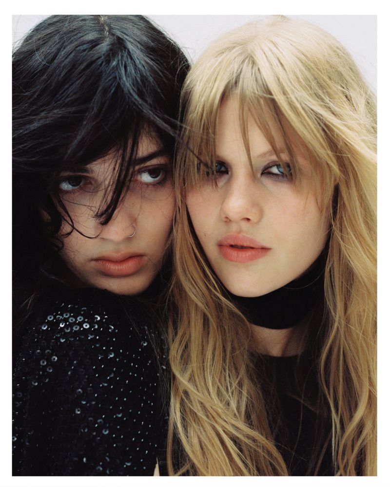 Close-up portrait of two female models with dark and blonde hair posing together against a plain white background.