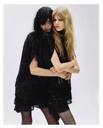 Two female models embracing closely while wearing black sequined dresses against a clean white seamless studio background.