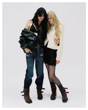 Two female models posing together against a plain white studio background wearing dark outerwear and studded jeans