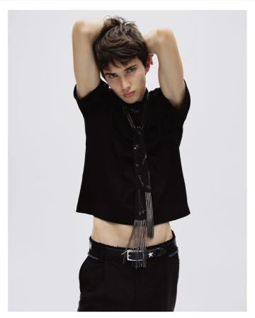 A young male model posing with his arms raised, wearing a black t-shirt and dark trousers against a white seamless backdrop.
