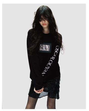 Model with dark hair wearing a black long-sleeve sweatshirt with white text and a textured skirt against a grey background.
