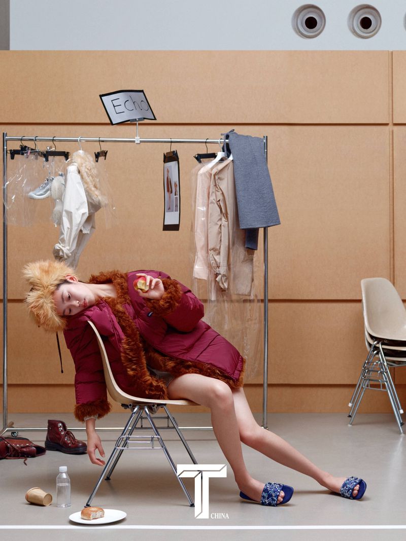 Model in a burgundy puffer jacket resting on a chair with clothing racks behind in a T Magazine China editorial by photographer Liu Song