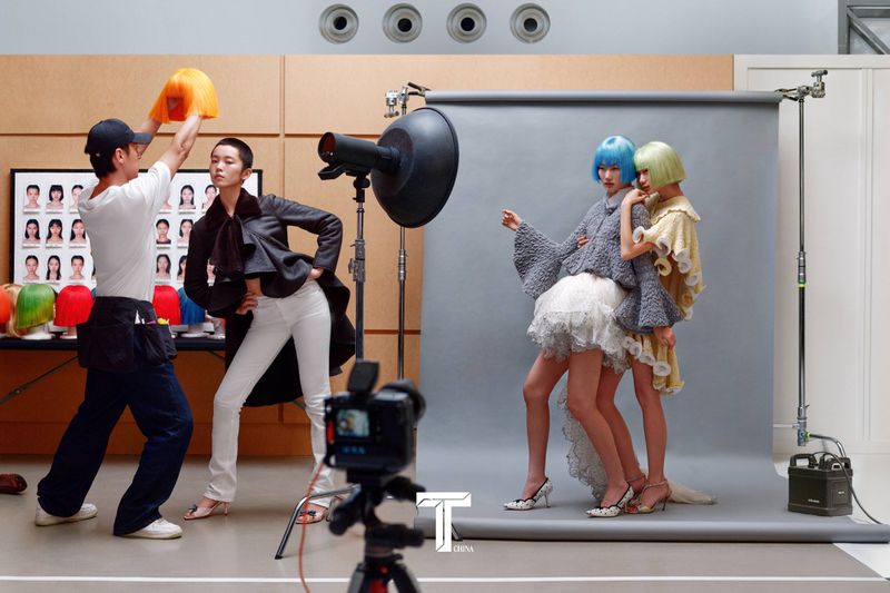 Behind-the-scenes fashion photo shoot with crew, studio lighting, and models wearing colorful wigs on a grey seamless backdrop
