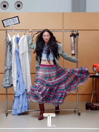 Model standing by a clothing rack wearing a ruffled plaid skirt, blue top, green blazer, and brown boots