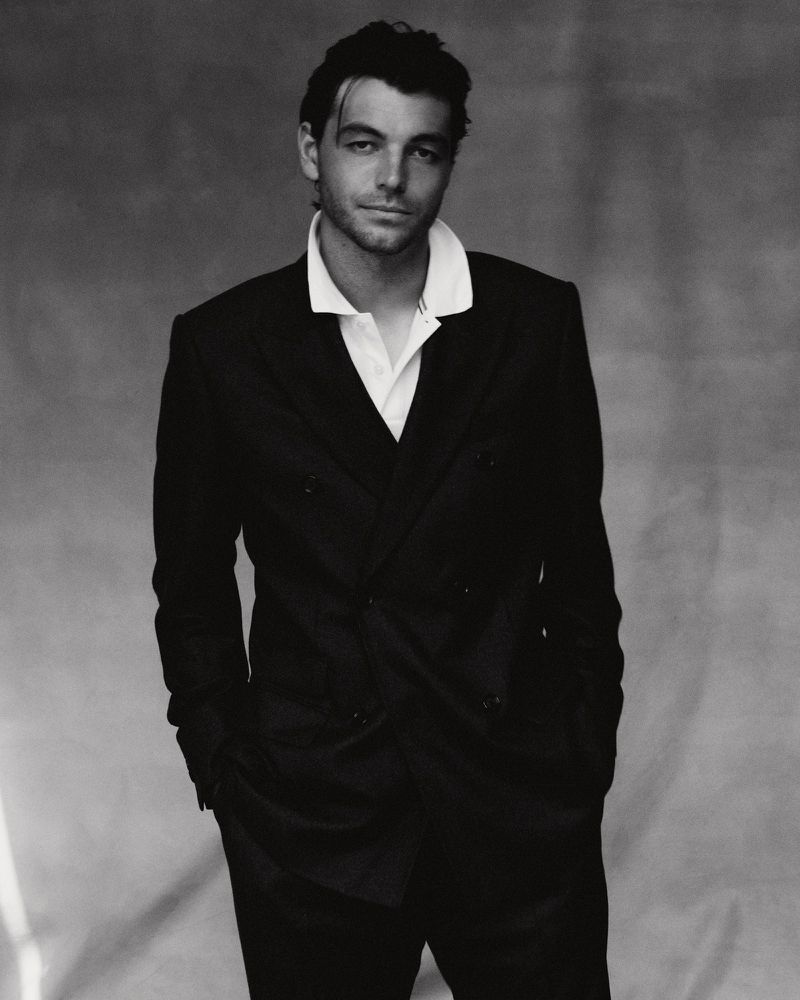 Portrait of Taylor Fritz wearing a dark double-breasted suit and white collared shirt against a textured grey backdrop.