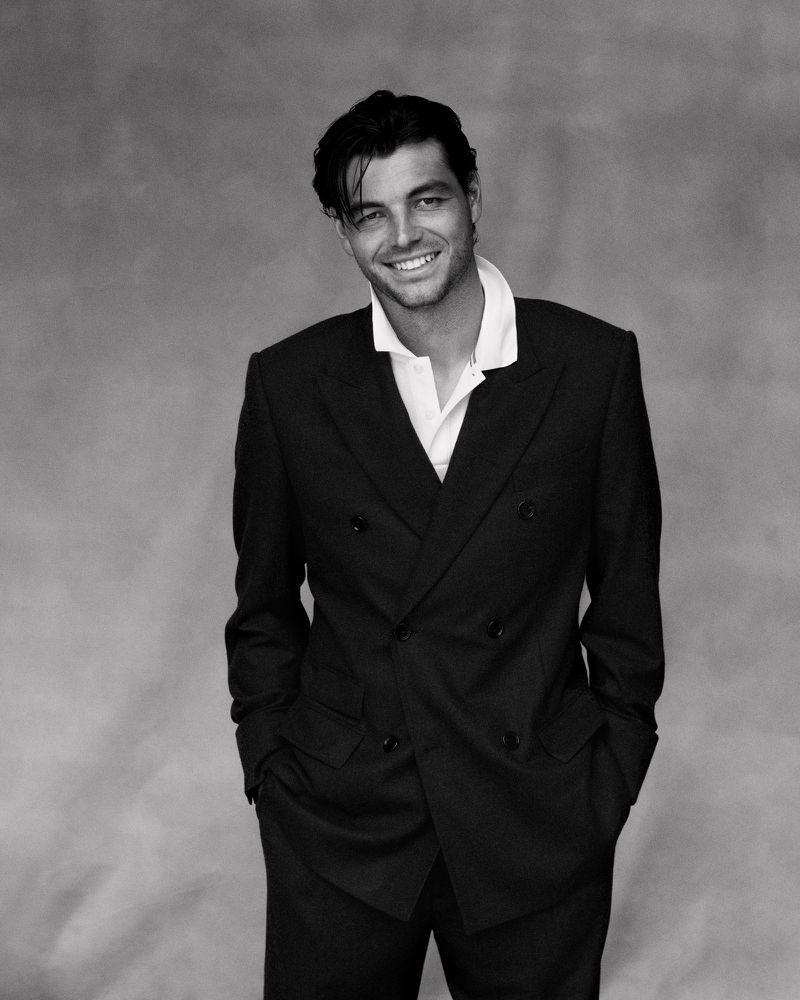 Portrait of Taylor Fritz wearing a double-breasted dark suit and white collared shirt against a grey textured studio backdrop.