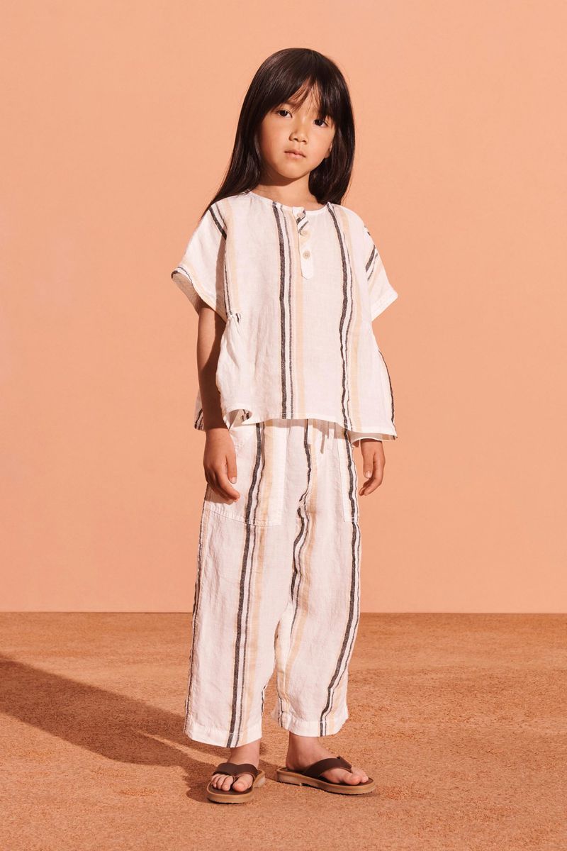 Full shot of an Asian child wearing a striped linen top and matching cropped pants, standing in a photo studio with a peach background.