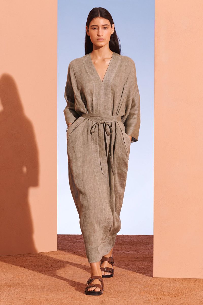 A woman in a taupe dress and dark brown sandals walks through an opening in a neutral-toned architectural setting with a blue sky background.