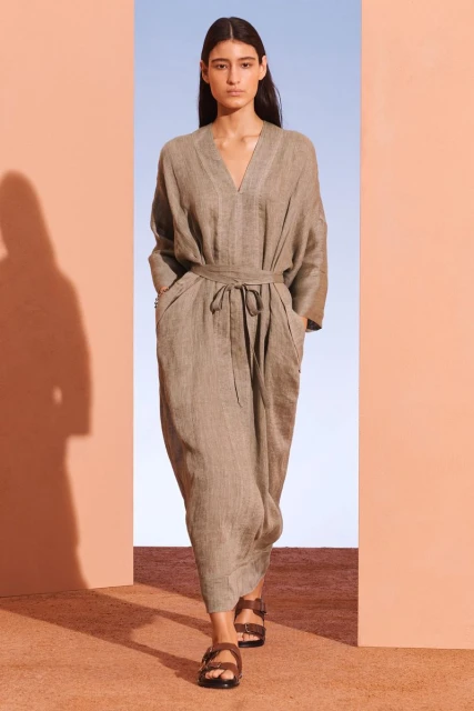 A woman in a taupe dress and dark brown sandals walks through an opening in a neutral-toned architectural setting with a blue sky background.