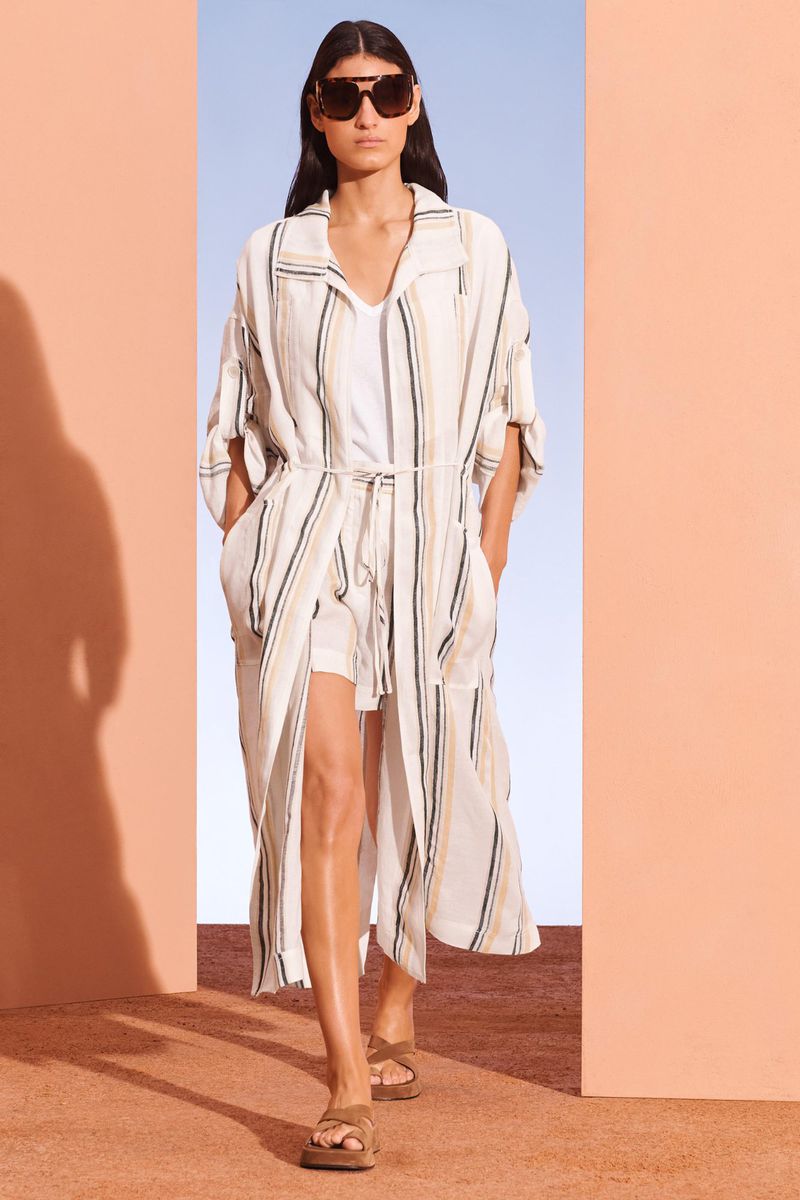 Fashion model showcases a striped duster coat, sunglasses, and sandals in a minimalist setting with peach and blue walls.