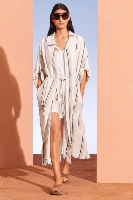 Fashion model showcases a striped duster coat, sunglasses, and sandals in a minimalist setting with peach and blue walls.