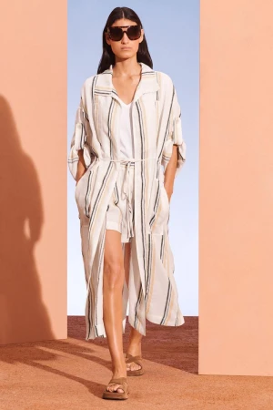 Fashion model showcases a striped duster coat, sunglasses, and sandals in a minimalist setting with peach and blue walls.
