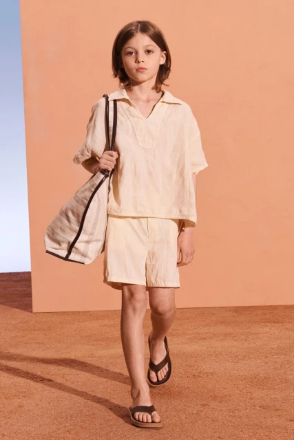 A child model in a light yellow matching shirt and shorts, carrying a tote bag and walking on a beige surface in a fashion editorial.