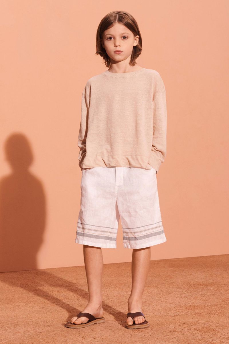 Young child in light casual summer fashion: tan top, white shorts with blue stripes, and dark sandals, poses indoors against peach wall.