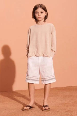 Young child in light casual summer fashion: tan top, white shorts with blue stripes, and dark sandals, poses indoors against peach wall.