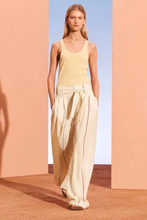 A fair-skinned model poses in a yellow tank top and striped palazzo pants against a backdrop divided into peach and blue sections.