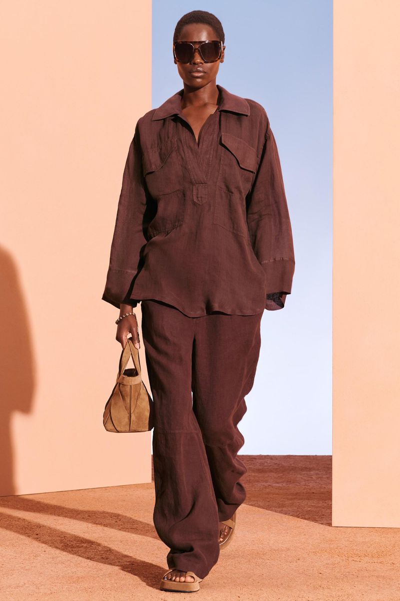 A black female fashion model wearing a brown long-sleeved shirt and wide-leg pants, holding a brown purse, walking on a brown runway.
