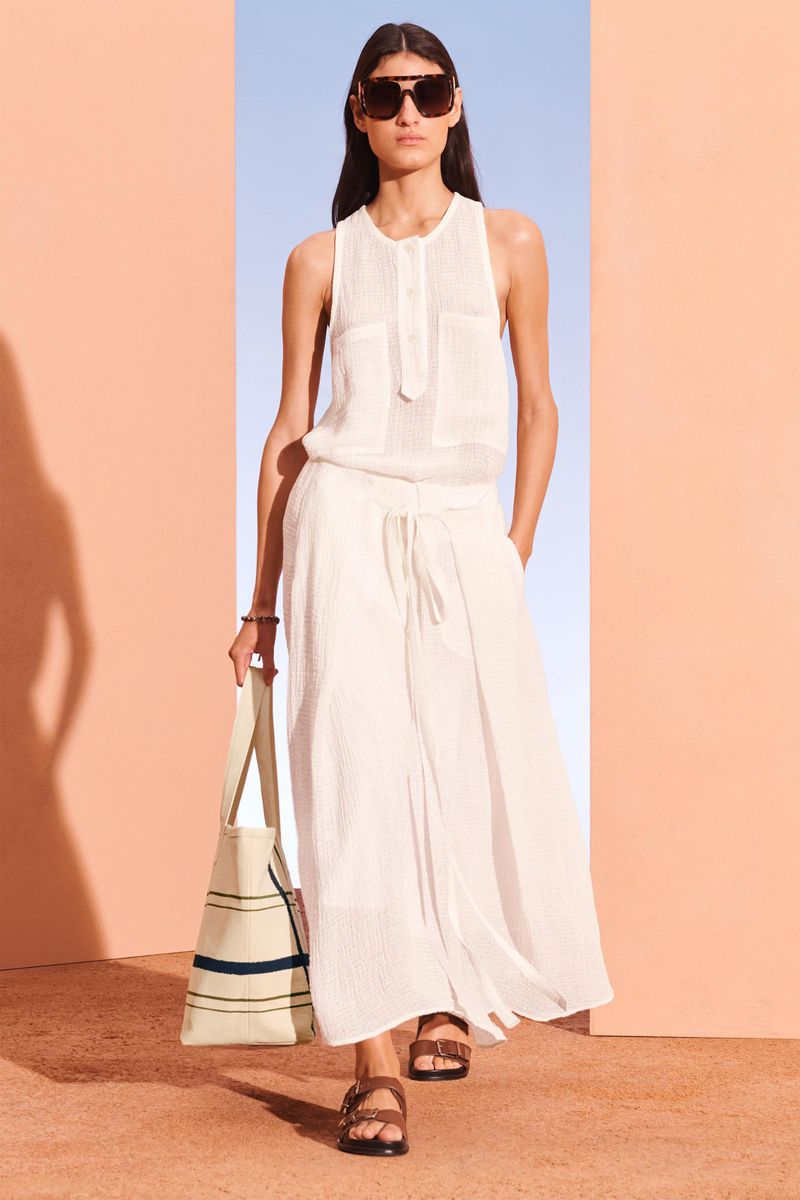 A woman in a white tank top and pants walks towards the camera, holding a striped tote bag, against a peach-and-blue background.