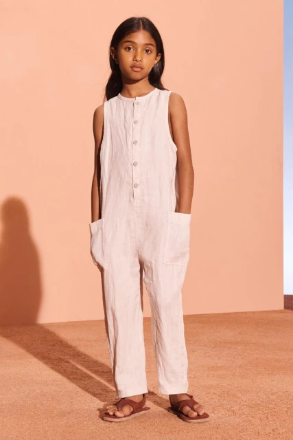 Portrait of a young girl wearing a sleeveless linen jumpsuit with brown sandals, posing in front of a coral and blue backdrop.