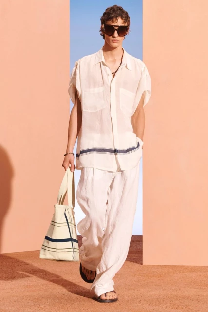 Male fashion model in a linen shirt and pants carrying a tote bag, posing against a light blue and peach backdrop in a studio setting.