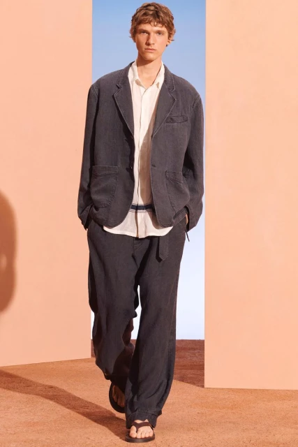 A male model in a linen suit walks in a brightly lit space with peach and blue walls. He wears dark sandals in this fashion shot.