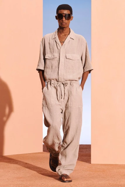 A black male fashion model showcases beige linen shirt and drawstring pants with sunglasses while walking indoors on a peach surface.