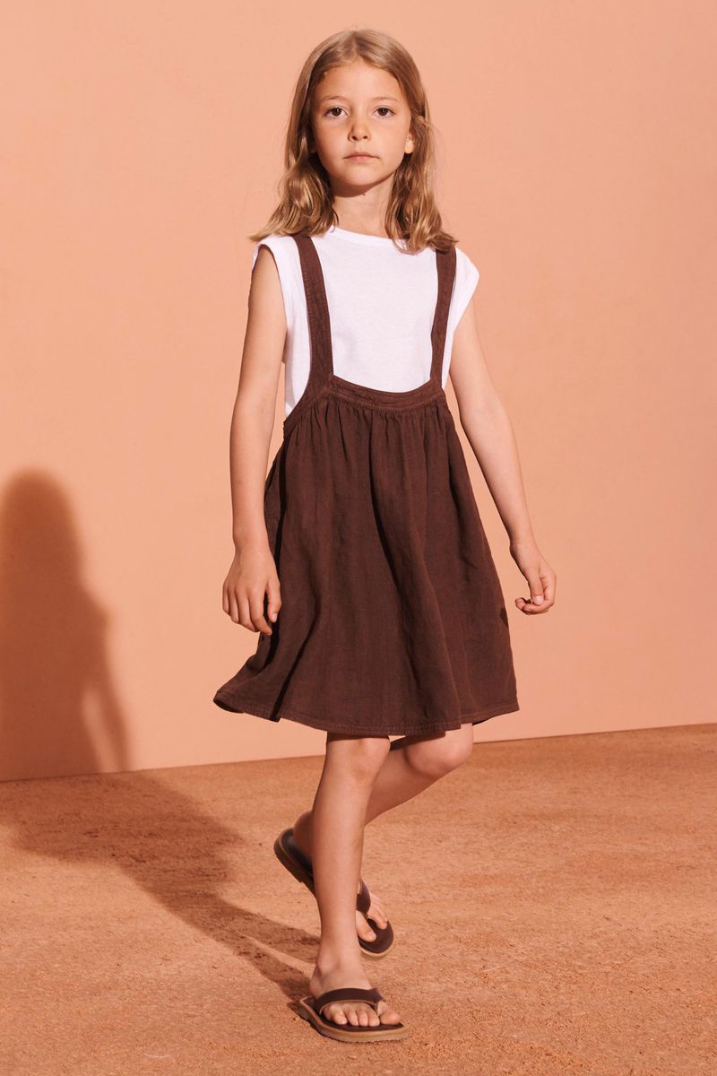 Young girl in a brown dress and white t-shirt walking on a brown surface, against a neutral peach-colored backdrop in a studio setting.