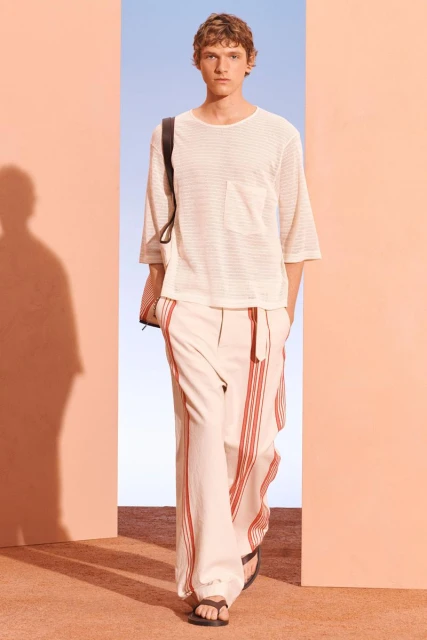 A male model presents a cream-colored knit t-shirt and striped trousers on a runway during a fashion show, in front of a peach and blue backdrop.