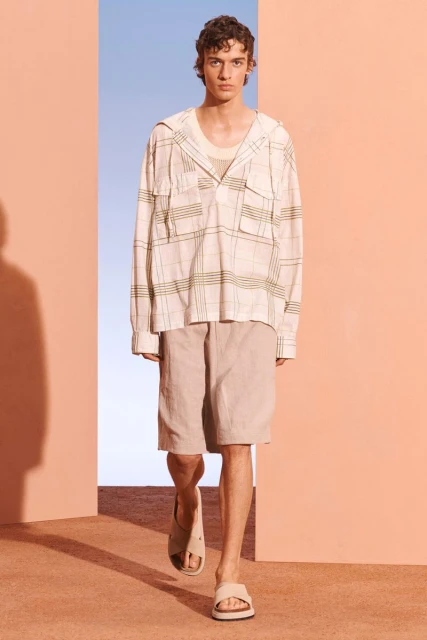 A young adult male fashion model walks toward the camera. He wears casual beige shorts, slide sandals, and a light plaid hooded shirt.